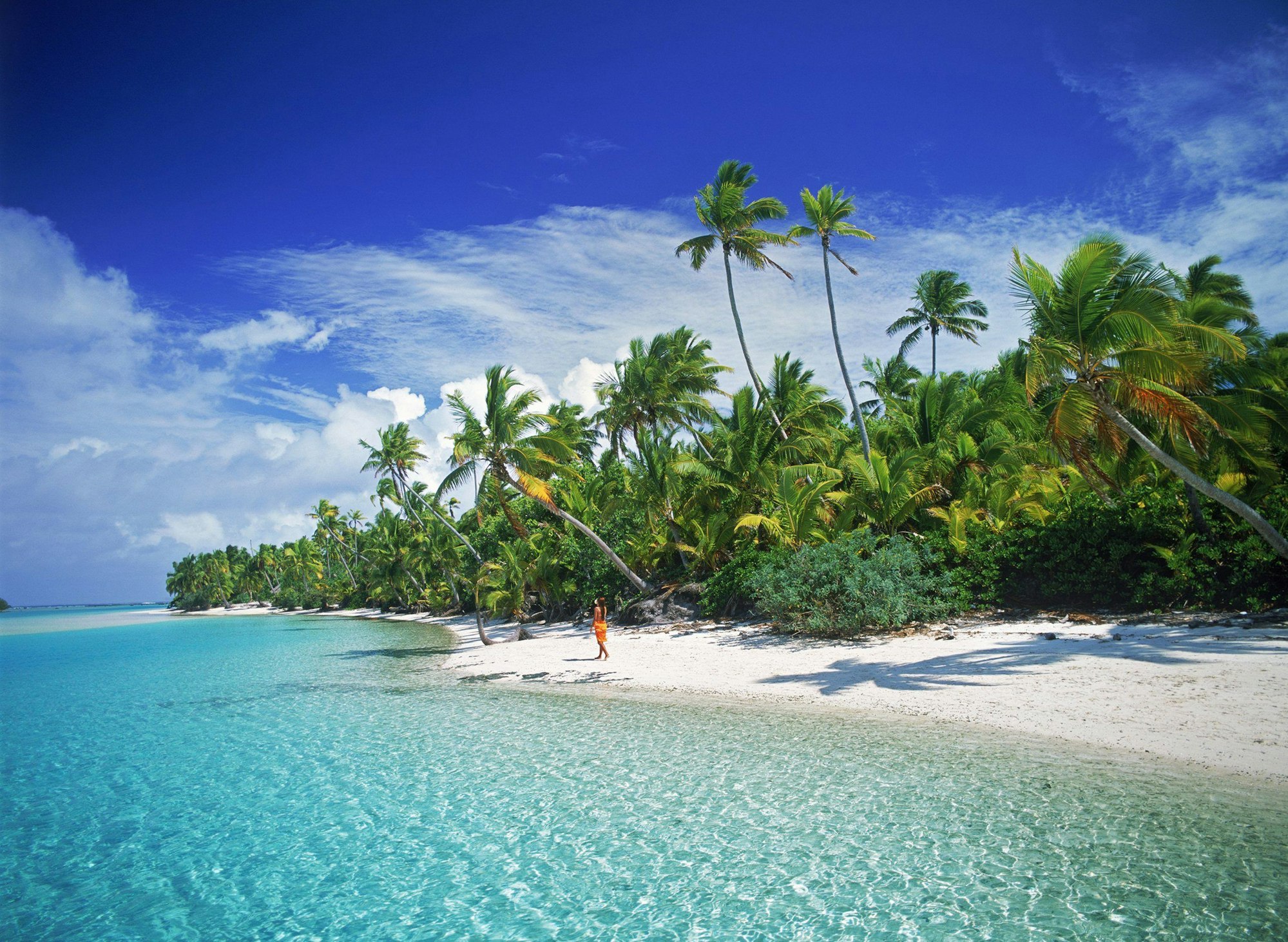 Hier zu sehen, eine Frau geht an einem Sandstrand spazieren. Links neben ihr befindet sich das türkise Meer, rechts befinden sich zahlreiche Palmen.