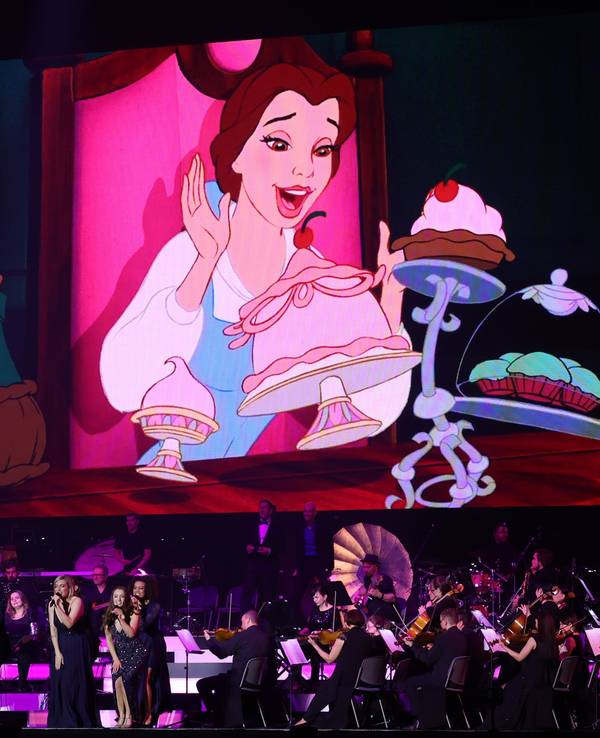 Das Orchester spielt auf der Bühne während man im Hintergrund eine Szene aus „Die Schöne und das Biest“ sieht.