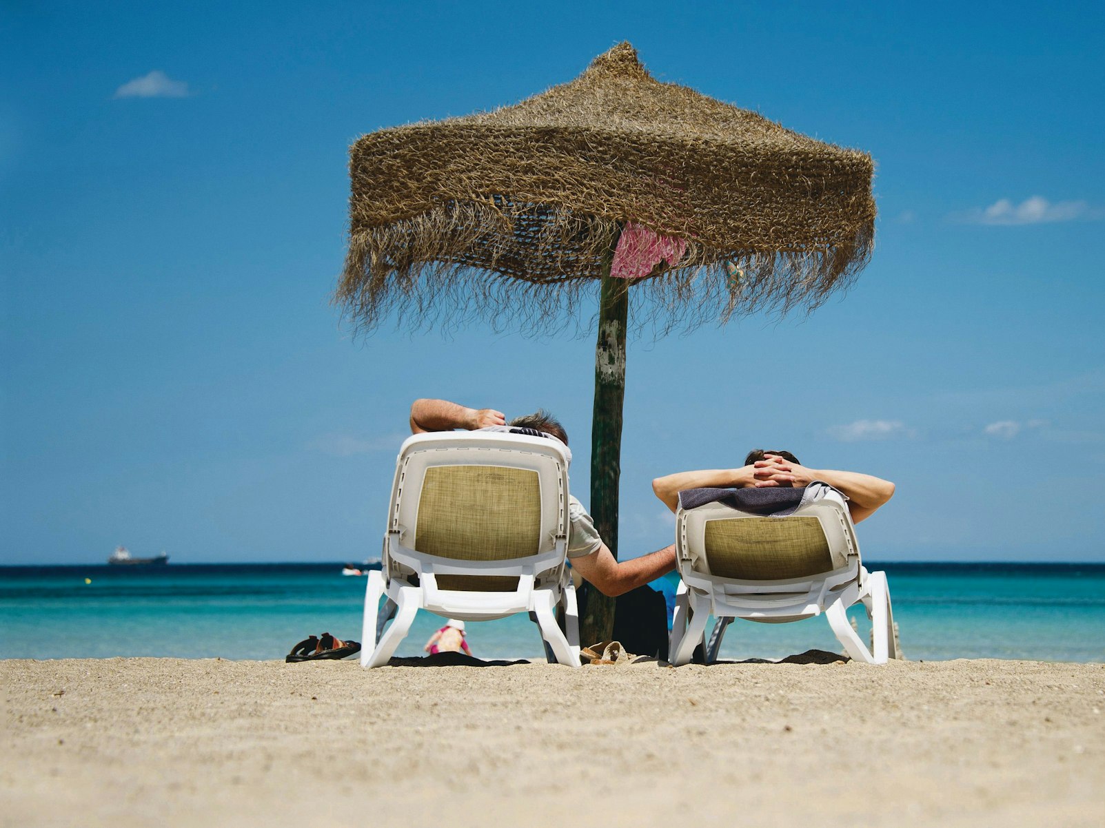 Zwei Urlauber auf Liegen unter einem Sonnenschirm an der Plaja de Muro auf Mallorca.