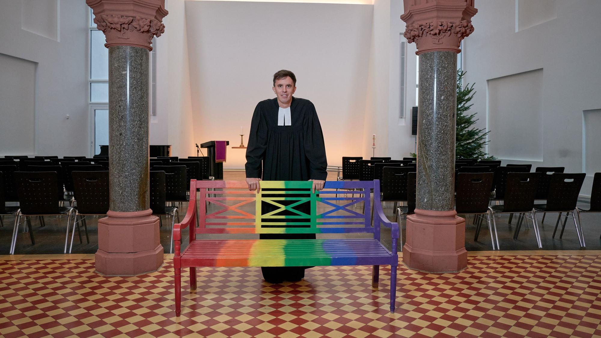 Pastor Tim Lahr steht an einer bunten Bank in der Christuskirche in Köln.