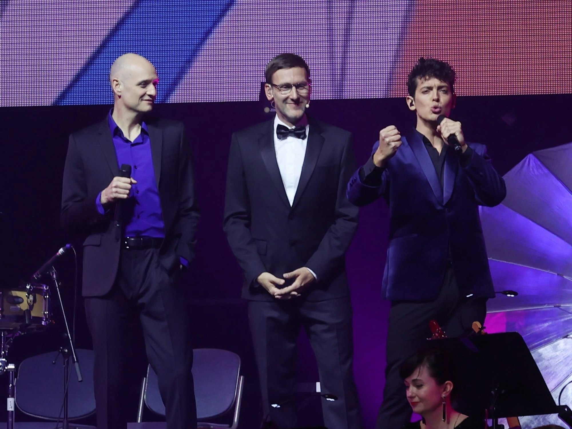 Auf der Bühne der Lanxess-Arena stehen Sänger Drew Sarich, Moderator Simon Beeck und Sänger Gonzalo Campos López.
