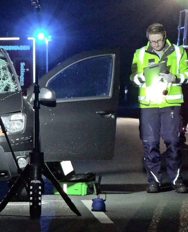 Scheinwerfer beleuchten auf einer Straße einen Pkw, dessen Frontscheibe gesplittert und Motorhaube eingedellt sind. Ein Polizist steht mit einem Klemmbrett daneben.