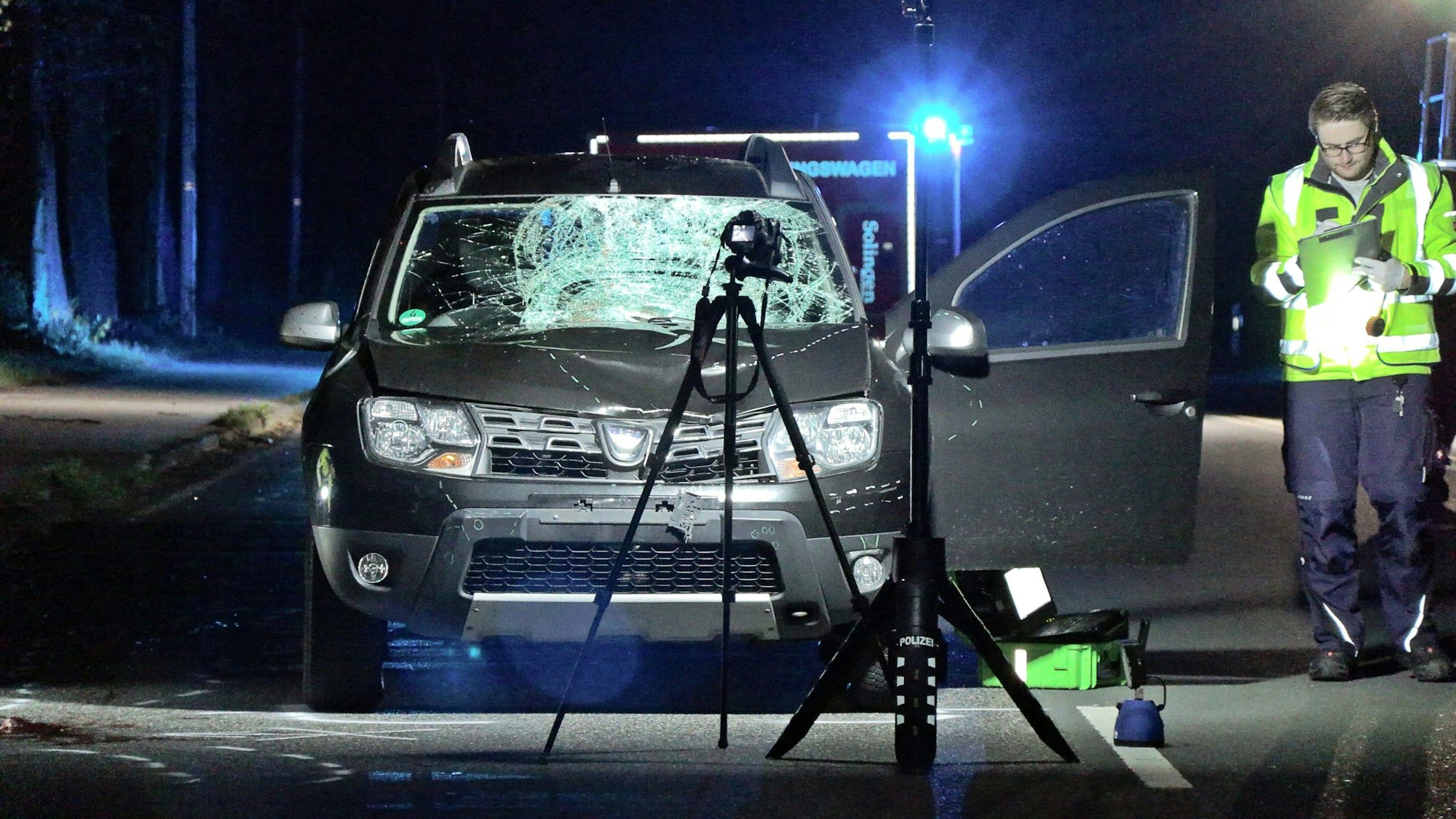 Scheinwerfer beleuchten auf einer Straße einen Pkw, dessen Frontscheibe gesplittert und Motorhaube eingedellt sind. Ein Polizist steht mit einem Klemmbrett daneben.
