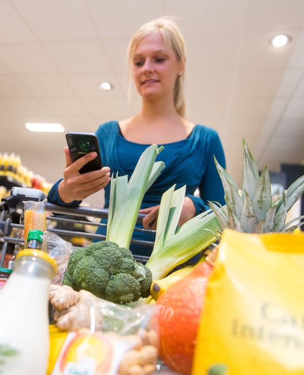 Eine Frau kauft in einem Supermarkt mit einem Einkaufswagen ein. In ihrer Hand hält sie ein Smartphone.