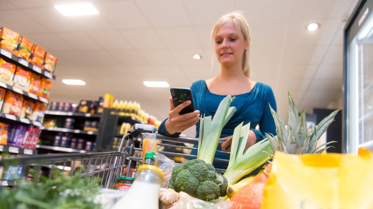 Eine Frau kauft in einem Supermarkt mit einem Einkaufswagen ein. In ihrer Hand hält sie ein Smartphone.