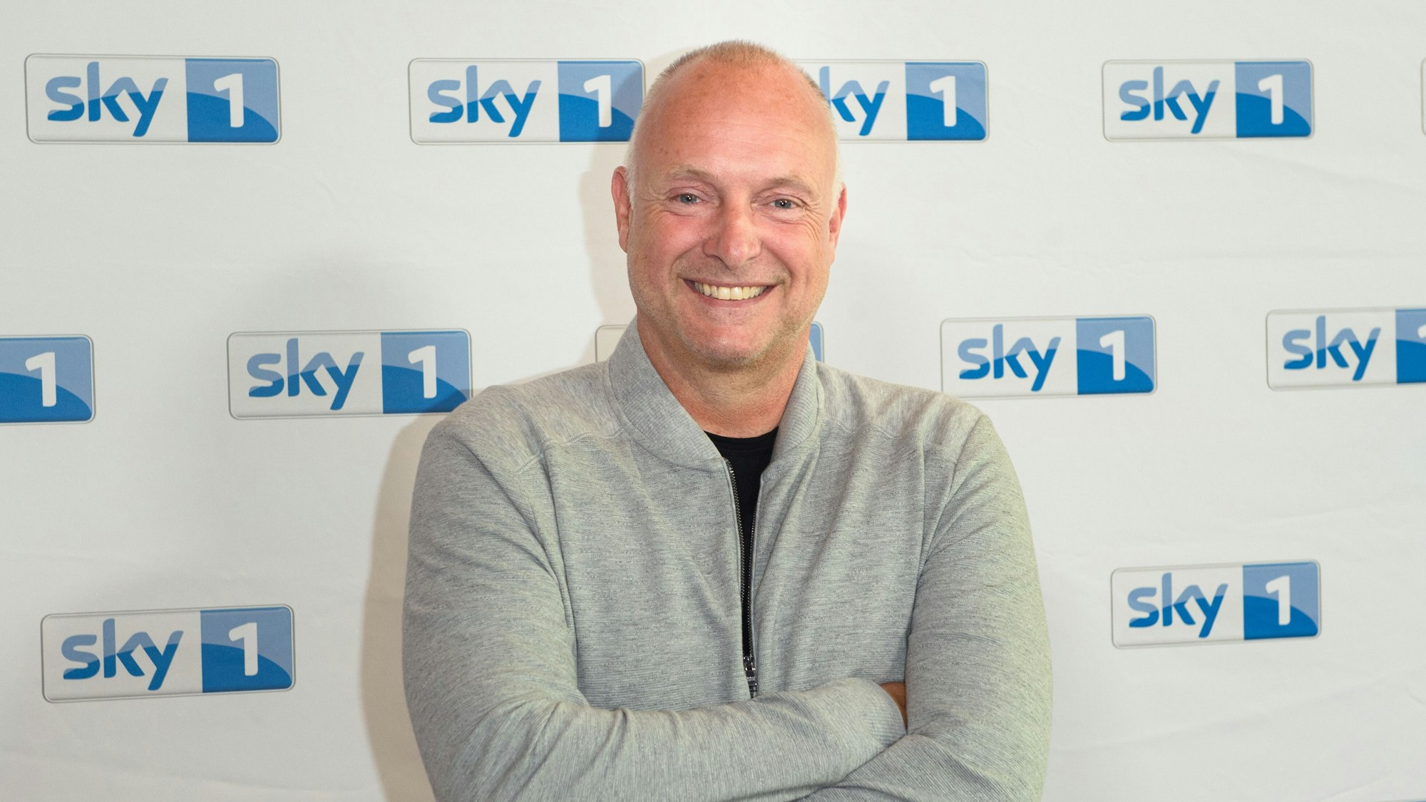 Frank Buschmann, Sportreporter und Fernsehmoderator, steht vor einer Logowand des Senders Sky