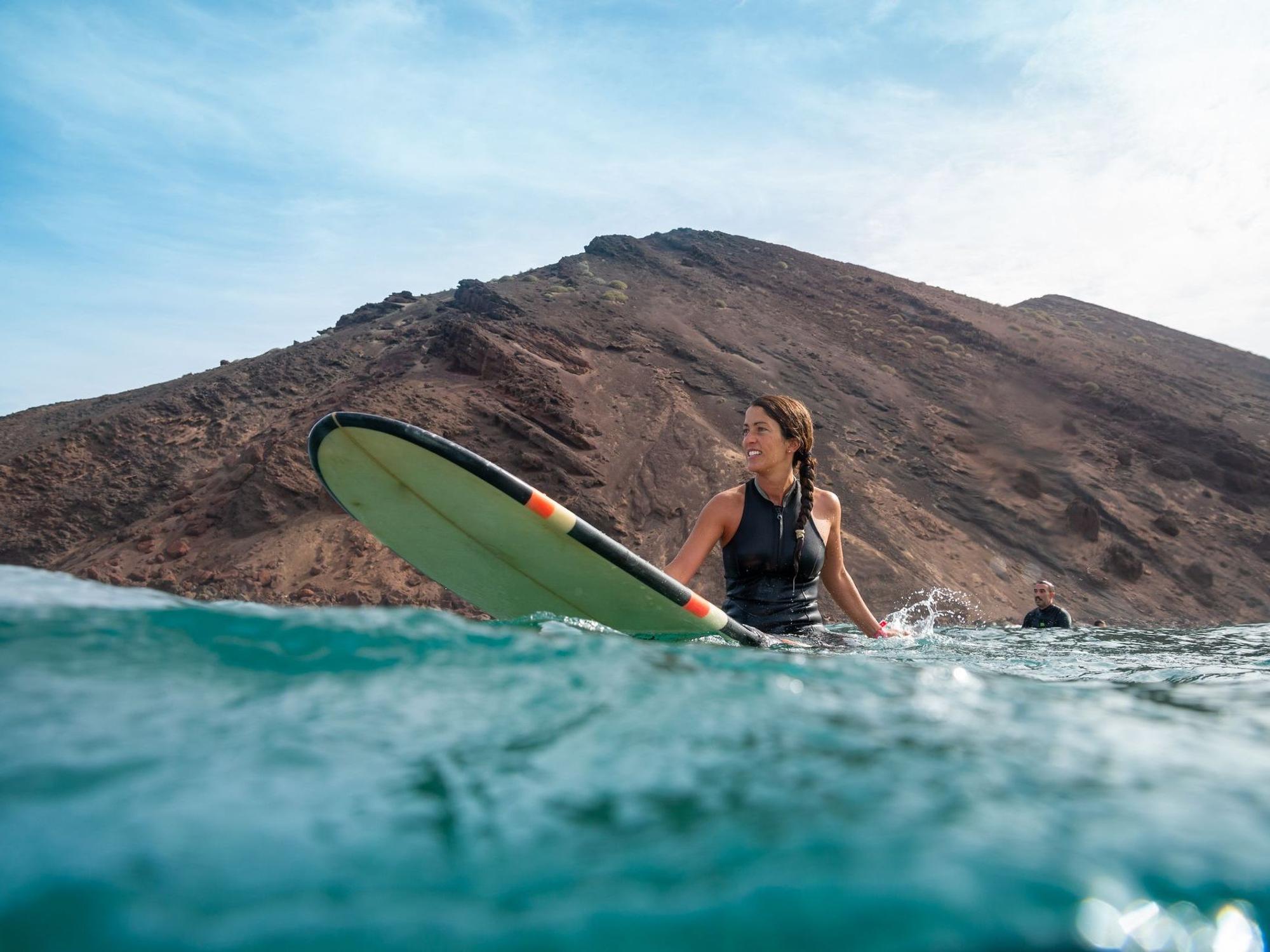 Eine brünette Frau sitzt auf ihrem Surfbrett im Wasser. Hinter ihr ein karger Berg und ein Mann.