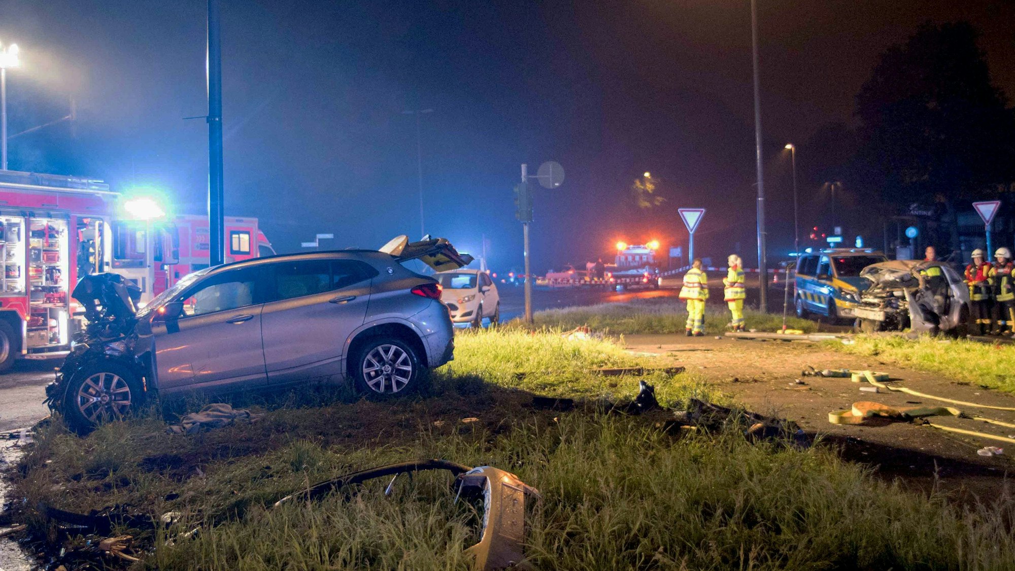 Zwei völlig beschädigte Autos stehen auf einer Grünfläche, dazwischen Einsatzkräfte von Feuerwehr und Polizei.
