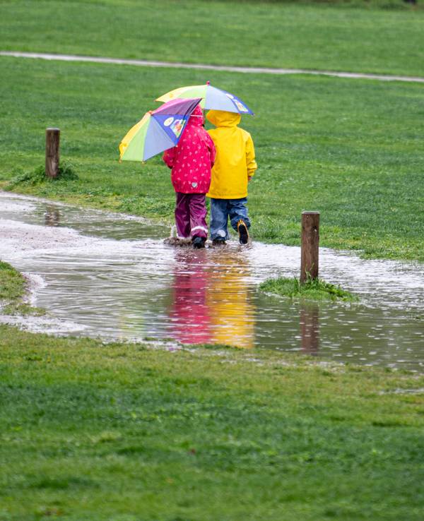 Zwei Kinder laufen in unserem Symbolbild einem Park durch eine große Pfütze. Im Westen Deutschlands lässt Starkregen die Flüsse steigen.