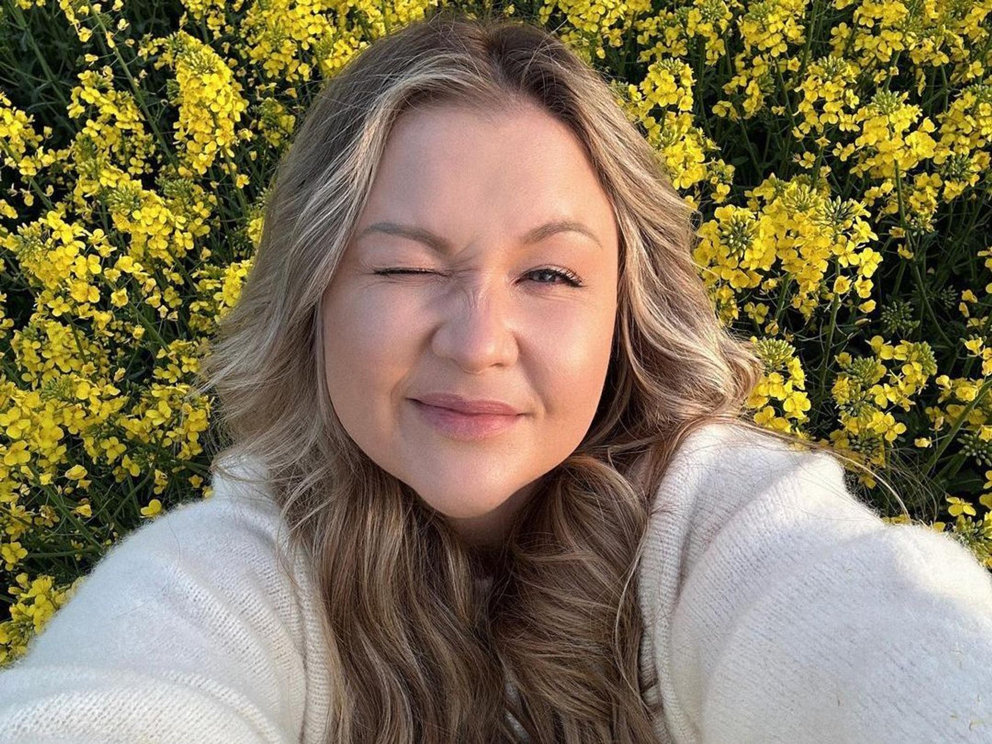 Sophie Thiel blickt inmitten eines gelben Rapsfeldes in die Kamera und zwinkert.