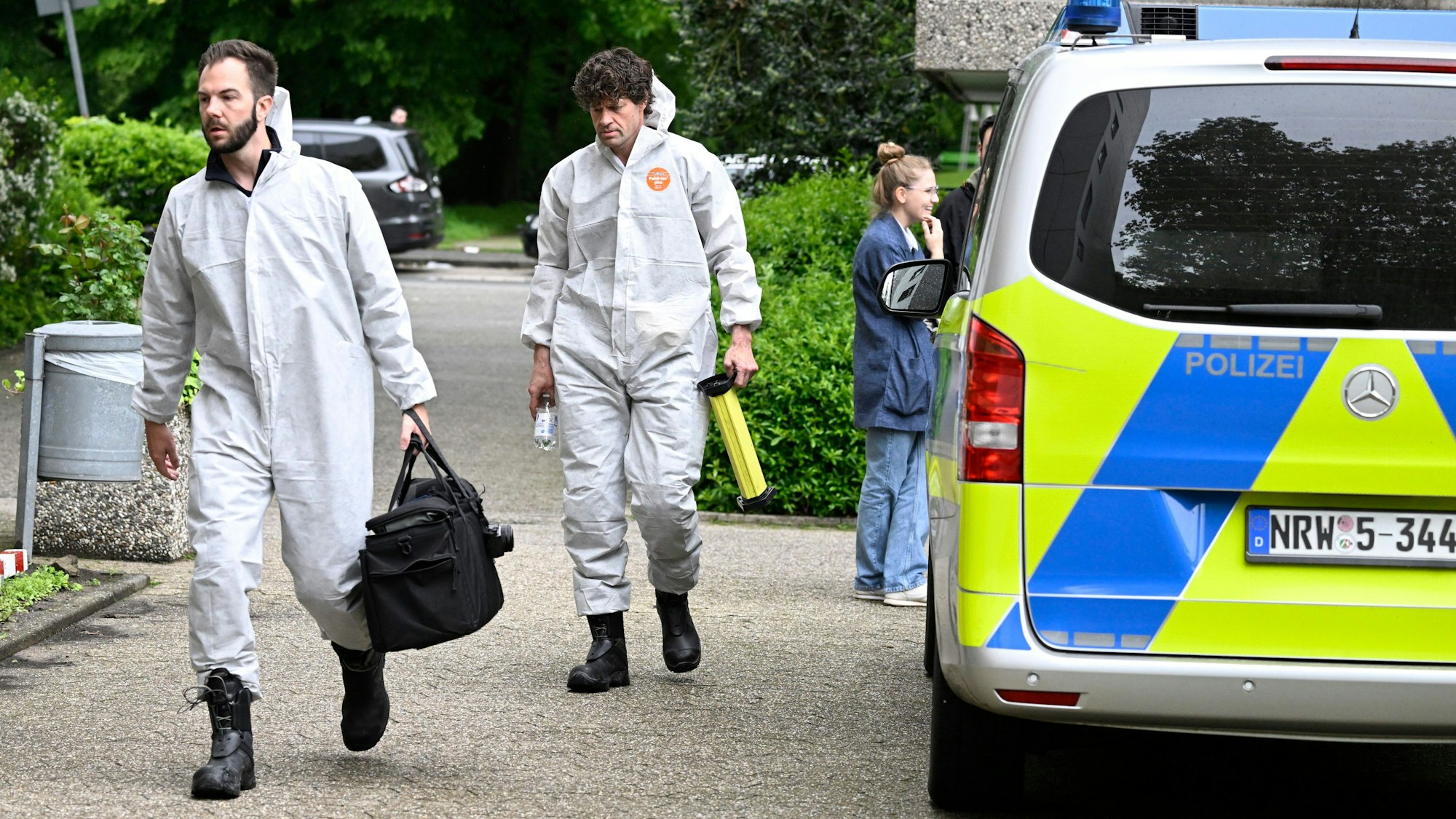 Zwei Männer in weißen Schutzanzügen gehen an einem Polizeifahrzeug vorbei.