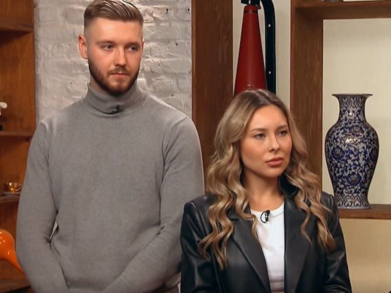 Das Paar Sarah Külper und Leopold Krey möchte eine Brosche verkaufen. Sarah trägt eine Lederjacke und ein weißes Oberteil. Er einen grauen Rollkragenpullover.