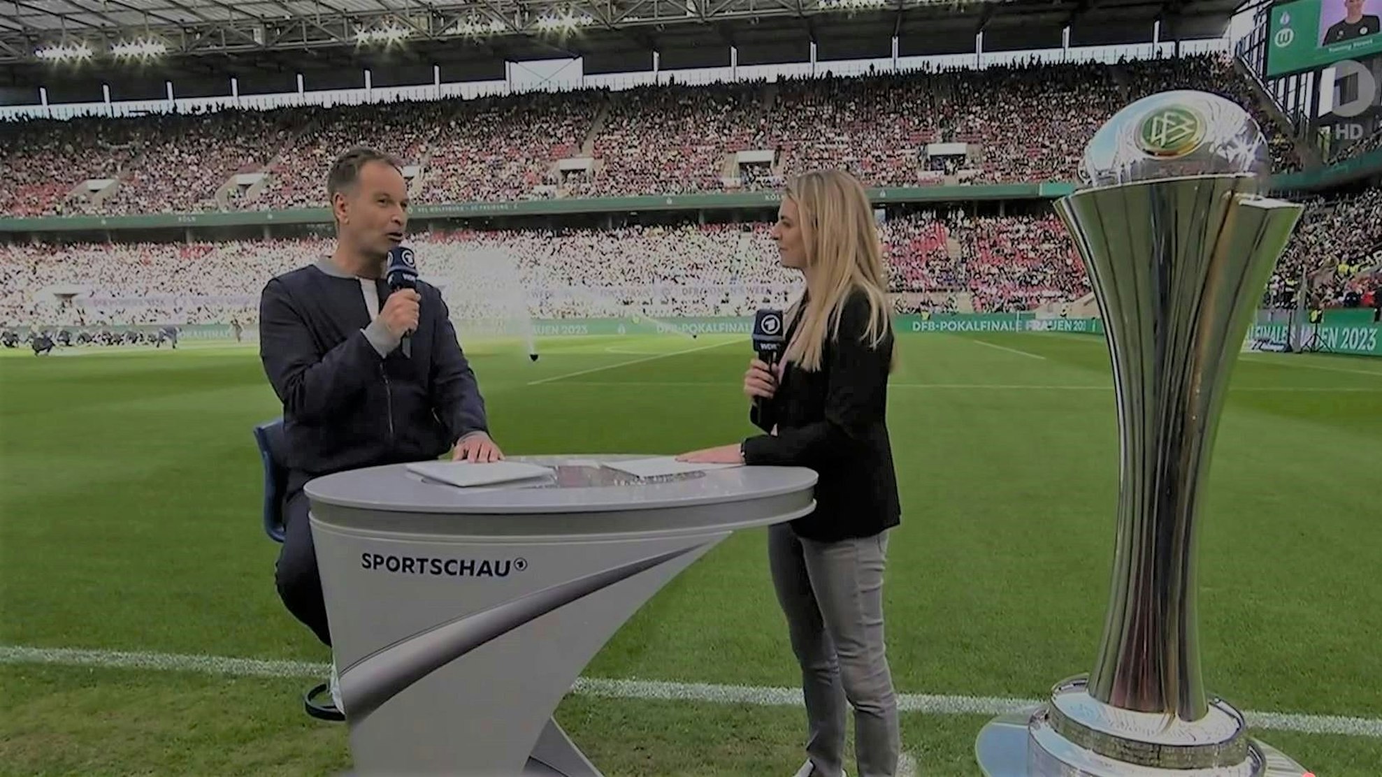Claus Lufen, sitzt auf einem Hocker, moderiert zusammen mit Nia Künzer im Rhein-Energie-Stadion.