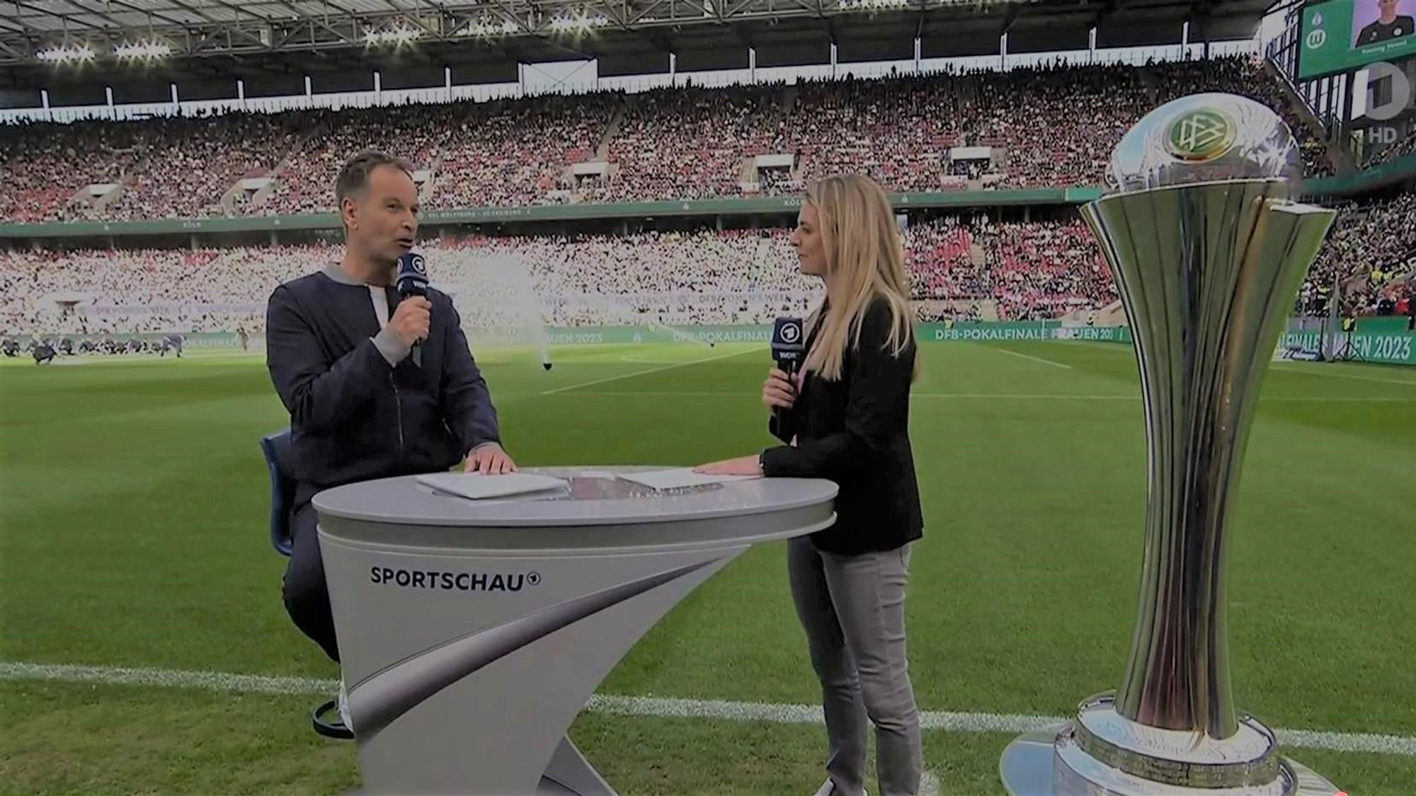 Claus Lufen, sitzt auf einem Hocker, moderiert zusammen mit Nia Künzer im Rhein-Energie-Stadion.
