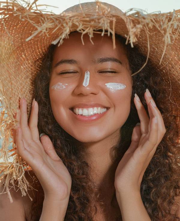 Lächelnde Frau mit Sonnensutzcreme im Gesicht und Strandhut auf dem Kopf.