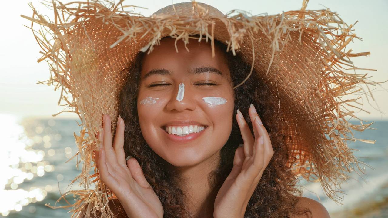 Lächelnde Frau mit Sonnensutzcreme im Gesicht und Strandhut auf dem Kopf.