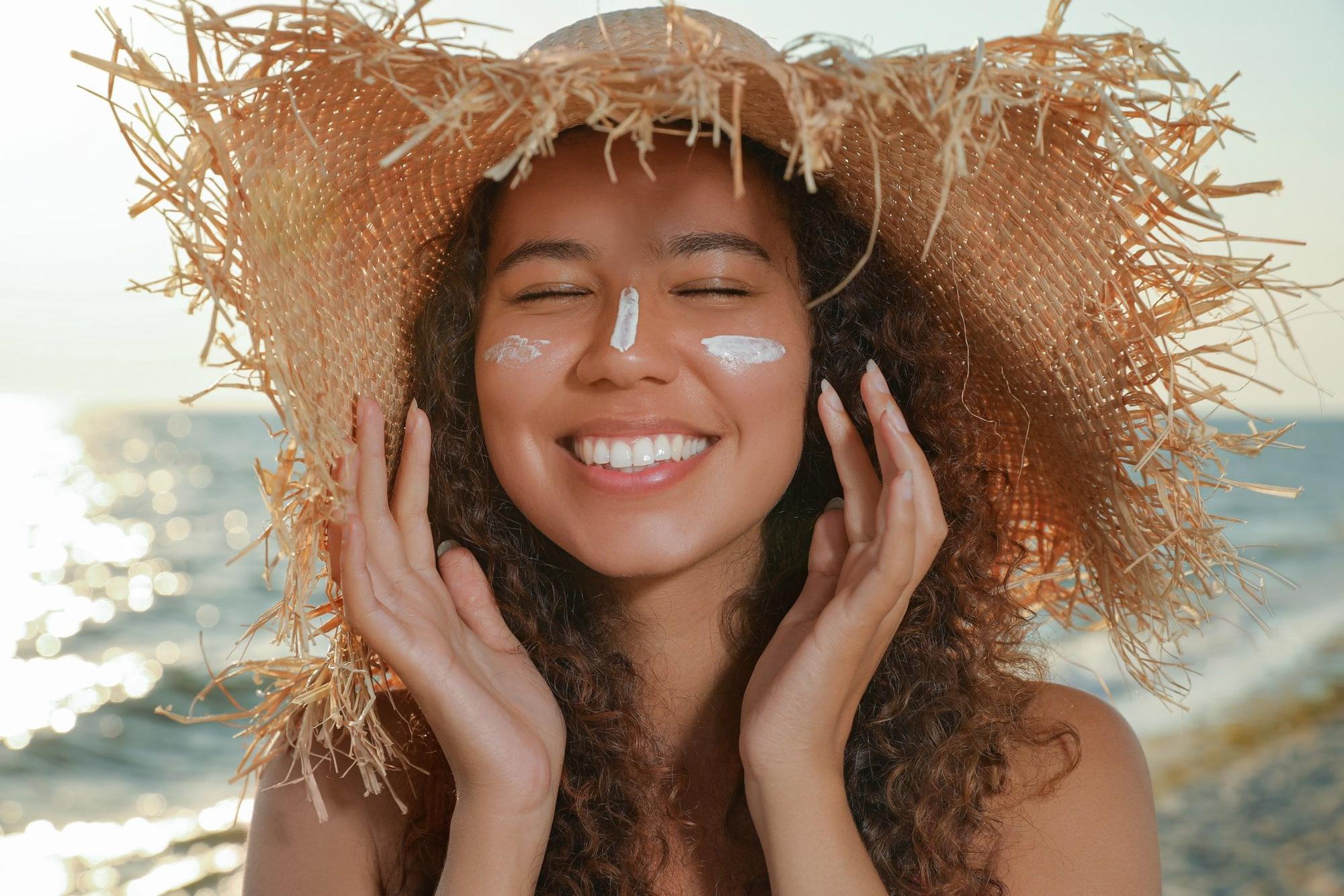 Lächelnde Frau mit Sonnensutzcreme im Gesicht und Strandhut auf dem Kopf.