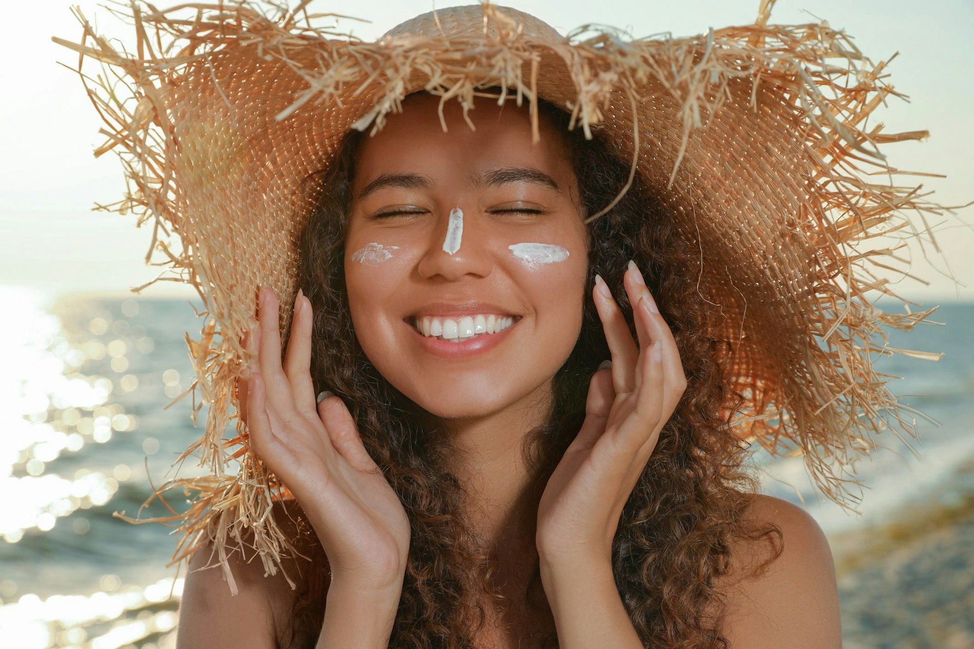 Lächelnde Frau mit Sonnensutzcreme im Gesicht und Strandhut auf dem Kopf.