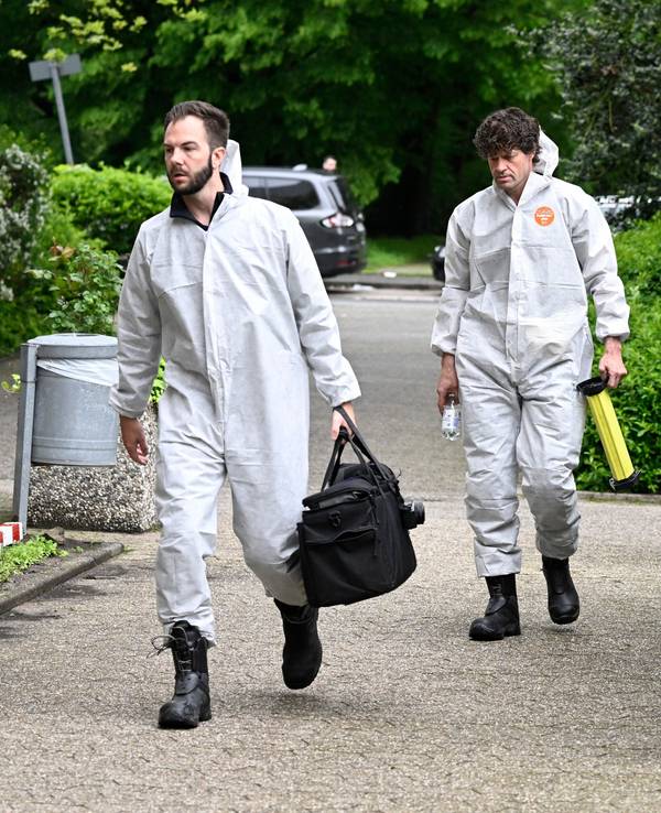 Zwei Beamte der Spurensicherung sind in Schutzanzügen auf dem Weg zu einem Tatort, dabei laufen sie an einem Polizeifahrzeug vorbei.