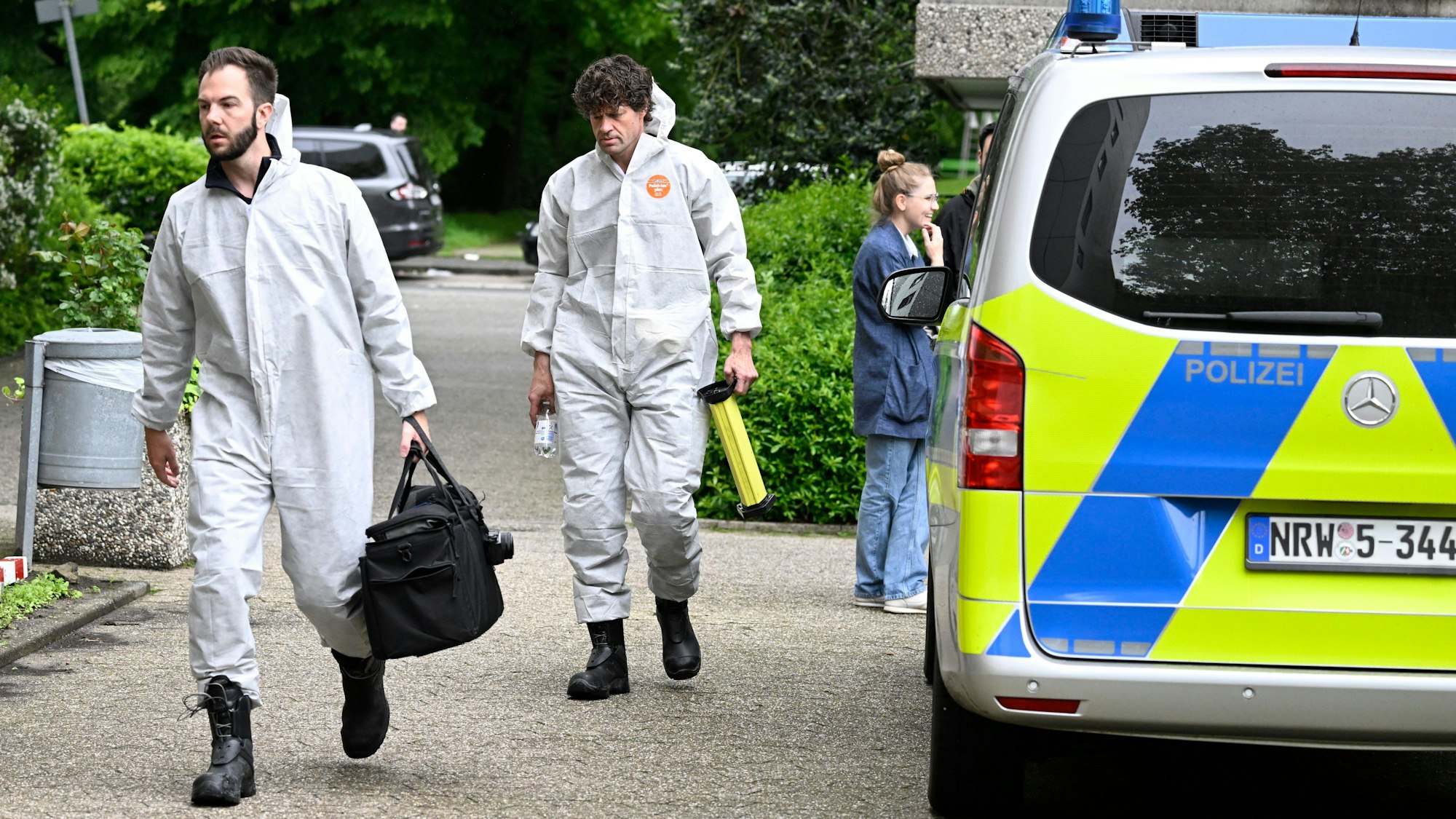 Zwei Beamte der Spurensicherung sind in Schutzanzügen auf dem Weg zu einem Tatort, dabei laufen sie an einem Polizeifahrzeug vorbei.