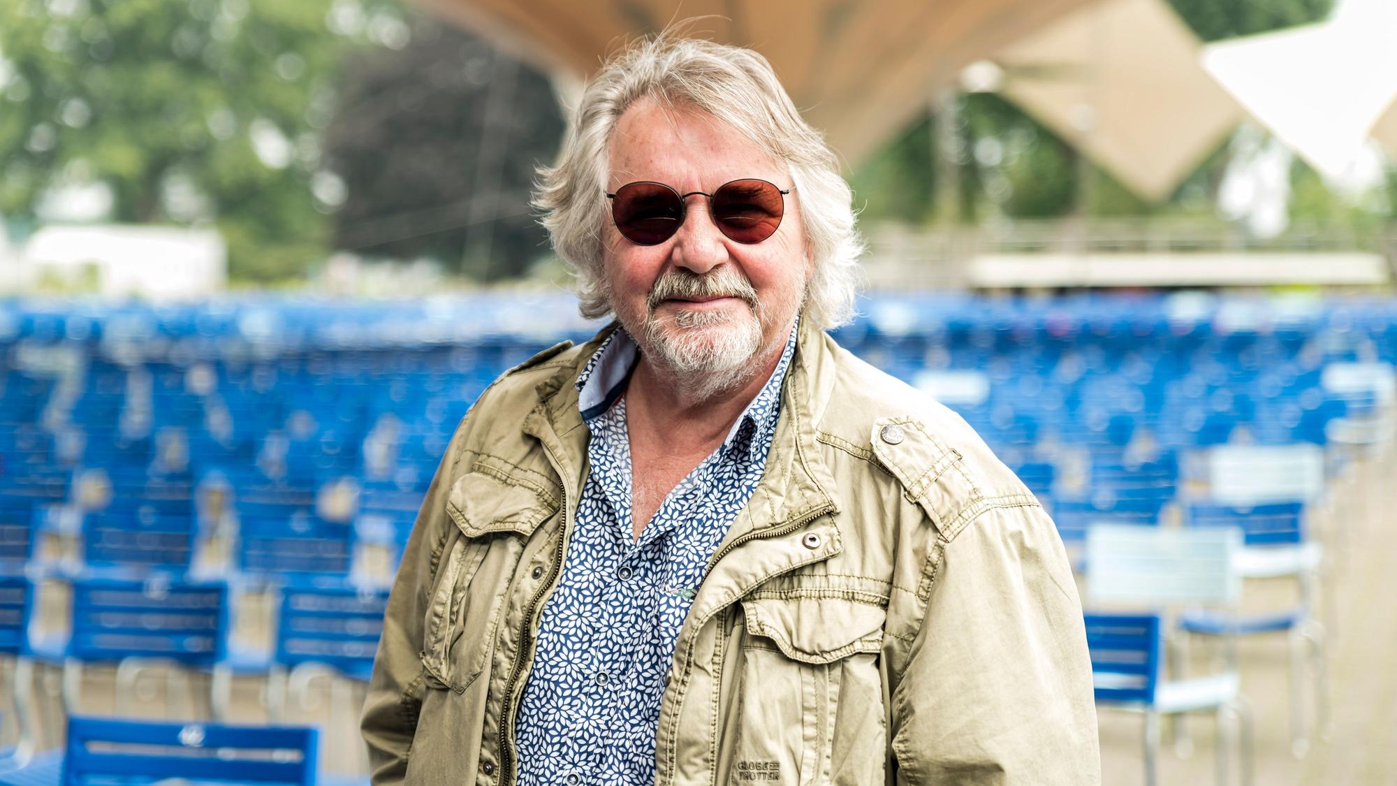 Tommy Engel steht mit Sonnenbrille im Tanzbrunnen vor leeren blauen Stühlen.