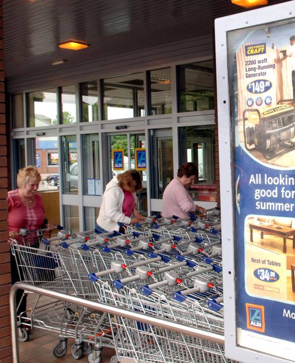 Aldi-Süd-Plakat mit Werbung an einer Filiale in England neben drei Frauen an Einkaufswagen