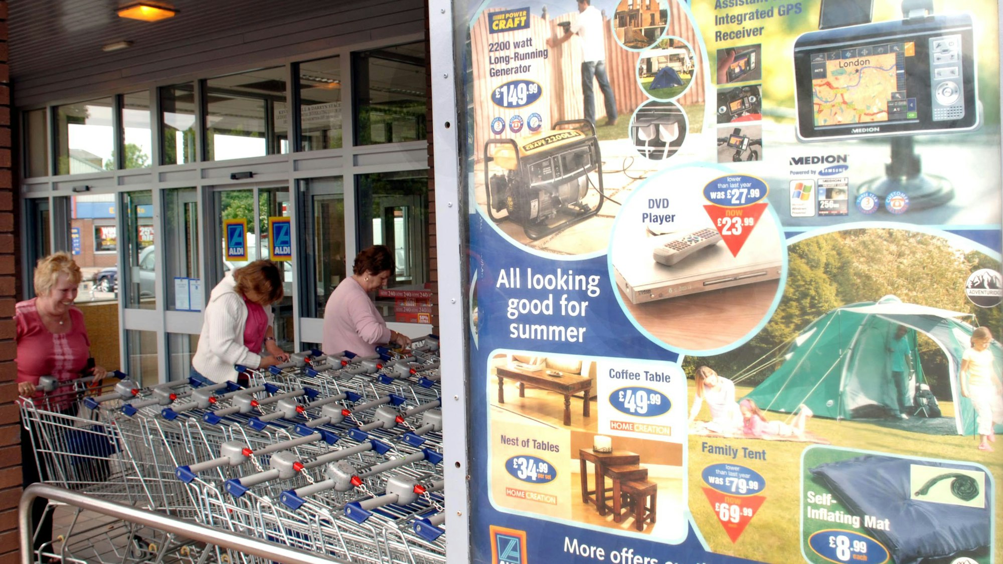 Aldi-Süd-Plakat mit Werbung an einer Filiale in England neben drei Frauen an Einkaufswagen