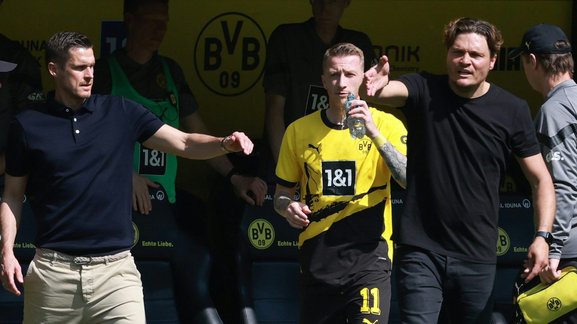 Sebastian Kehl, Marco Reus und Edin Terzic stehen an der Seitenlinie. Reus trinkt aus einer Wasserflasche, Kehl und Terzic gestikulieren.