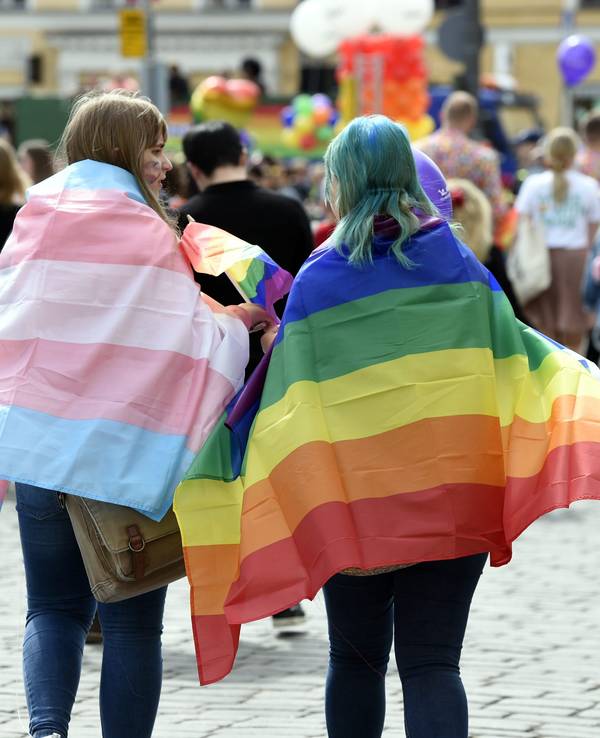 Zwei Personen mit einer Regenbogenflagge und einer Trans*-Flagge auf einer Parade.