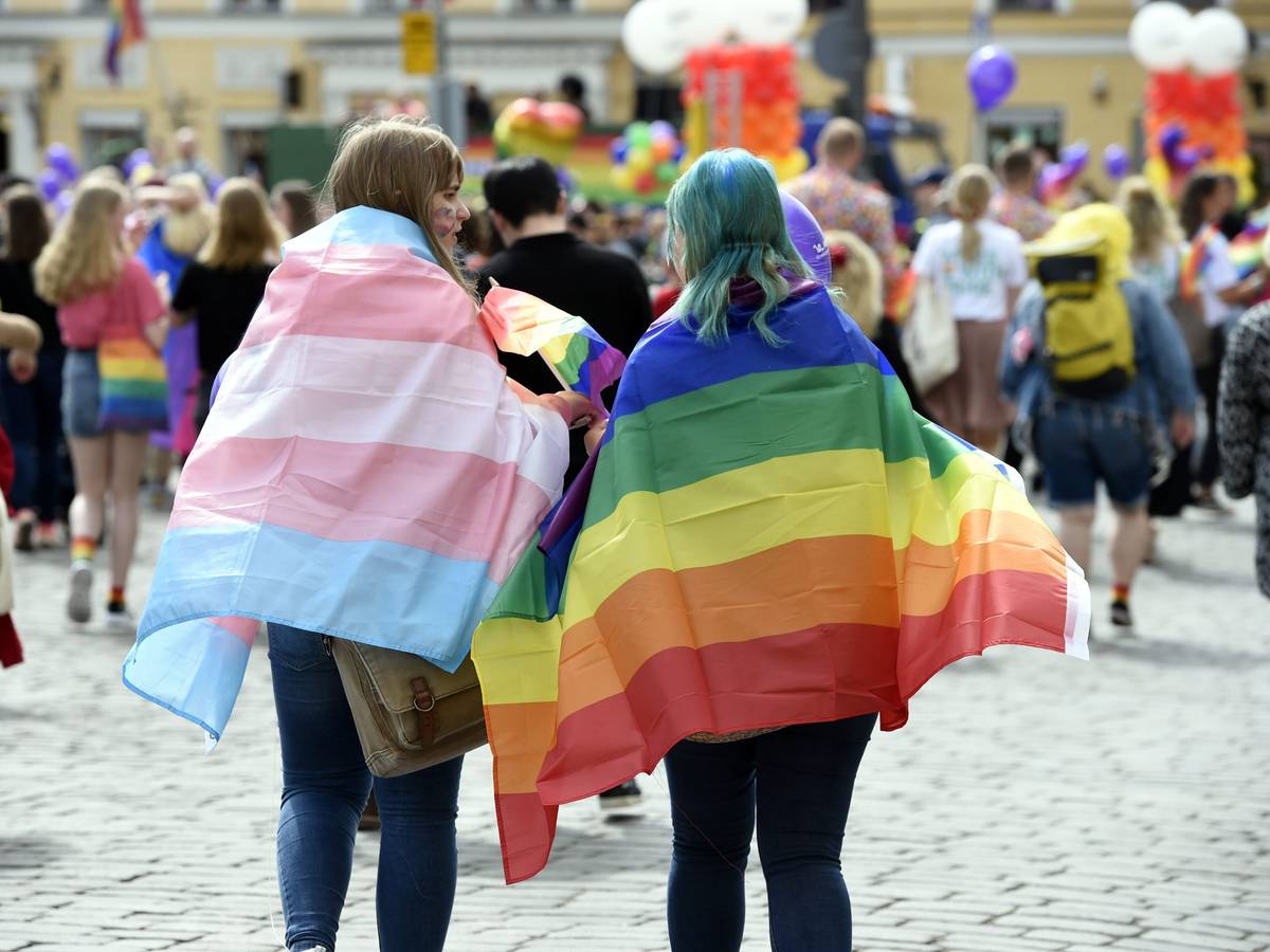 Zwei Personen mit einer Regenbogenflagge und einer Trans*-Flagge auf einer Parade.