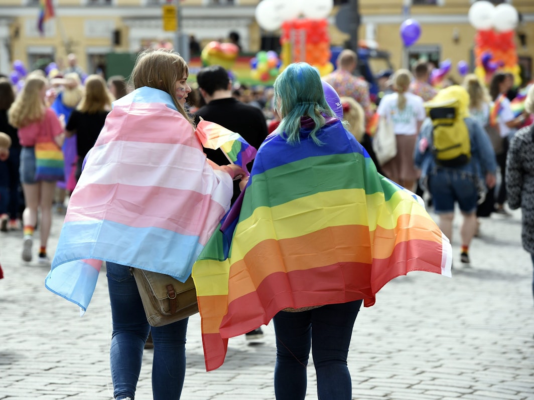 Zwei Personen mit einer Regenbogenflagge und einer Trans*-Flagge auf einer Parade.