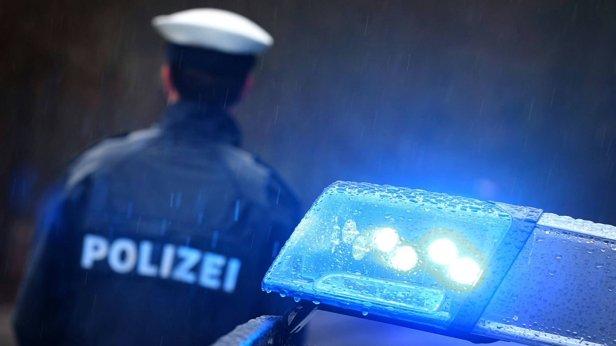 Ein Polizist steht im Regen vor einem Streifenwagen, dessen Blaulicht aktiviert ist.