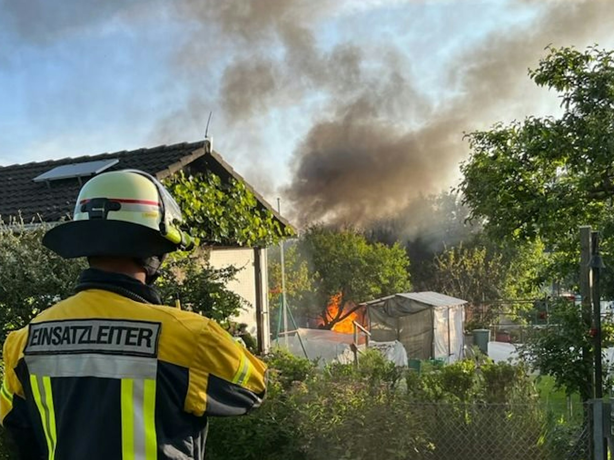 Ein Feuerwehrmann steht vor einer Gartenlaube, hinter der irgendetwas brennt.