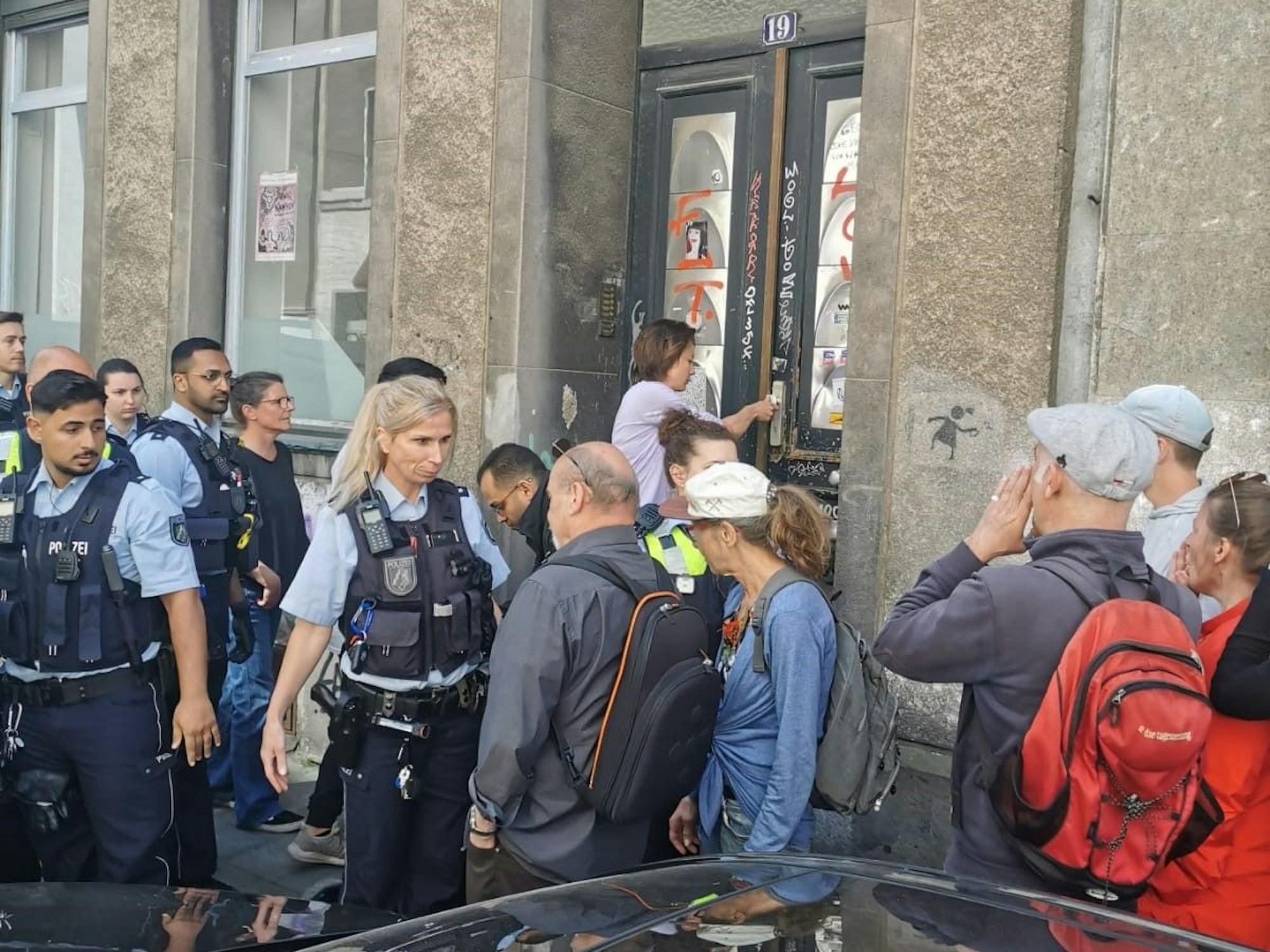 Mehrere Einsatzkräfte der Polizei stehen vor der Tür des Hauses, eine Beamtin unterhält sich mit einem Mann.