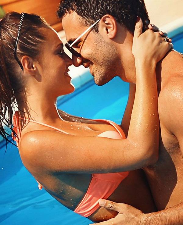 Frau und Mann in Badekleidung stehen im Swimming Pool, umarmen sich und schauen sich lächelnd in die Augen.