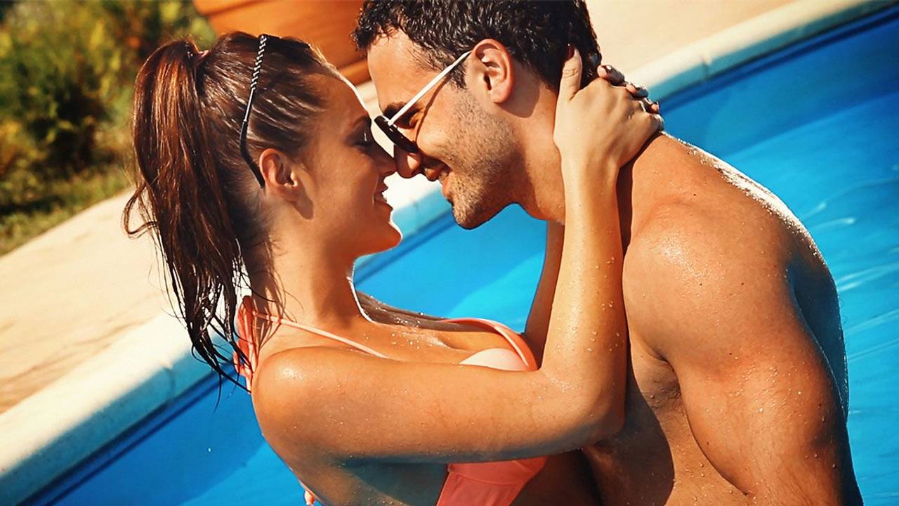 Frau und Mann in Badekleidung stehen im Swimming Pool, umarmen sich und schauen sich lächelnd in die Augen.