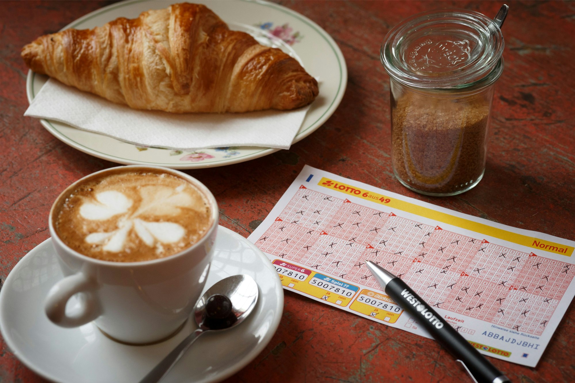 Lotto 6aus49 Spielschein auf Tisch neben Kaffeetasse und Croissant.