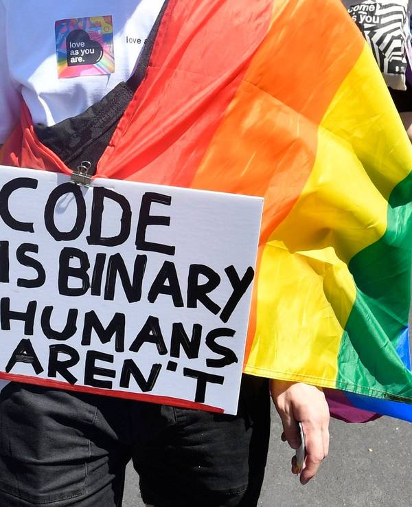 Demonstrierende Person mit einem beschrifteten Schild und einer Regenbogenflagge.