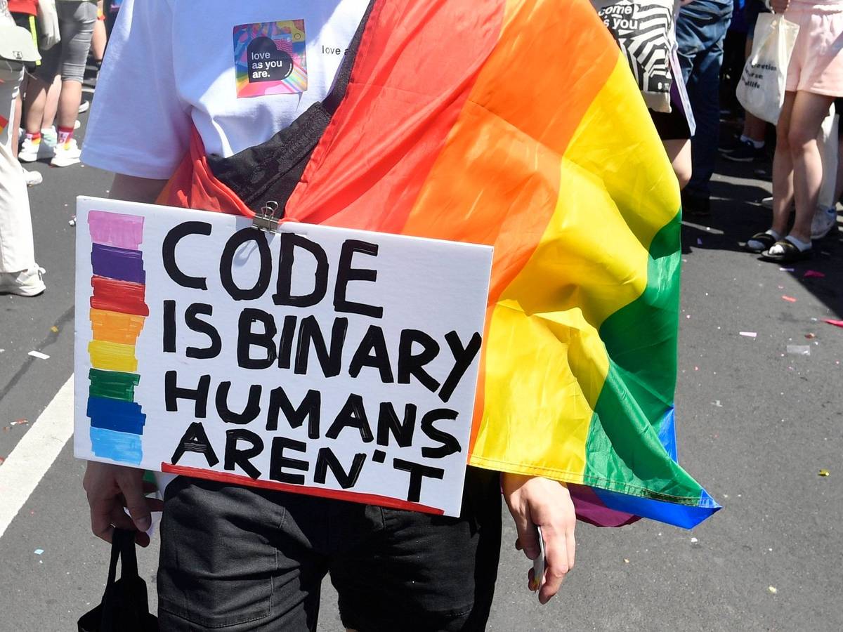 Demonstrierende Person mit einem beschrifteten Schild und einer Regenbogenflagge.