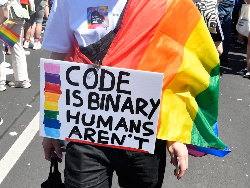 Demonstrierende Person mit einem beschrifteten Schild und einer Regenbogenflagge.