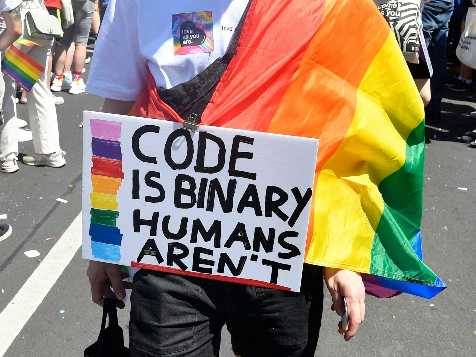 Demonstrierende Person mit einem beschrifteten Schild und einer Regenbogenflagge.