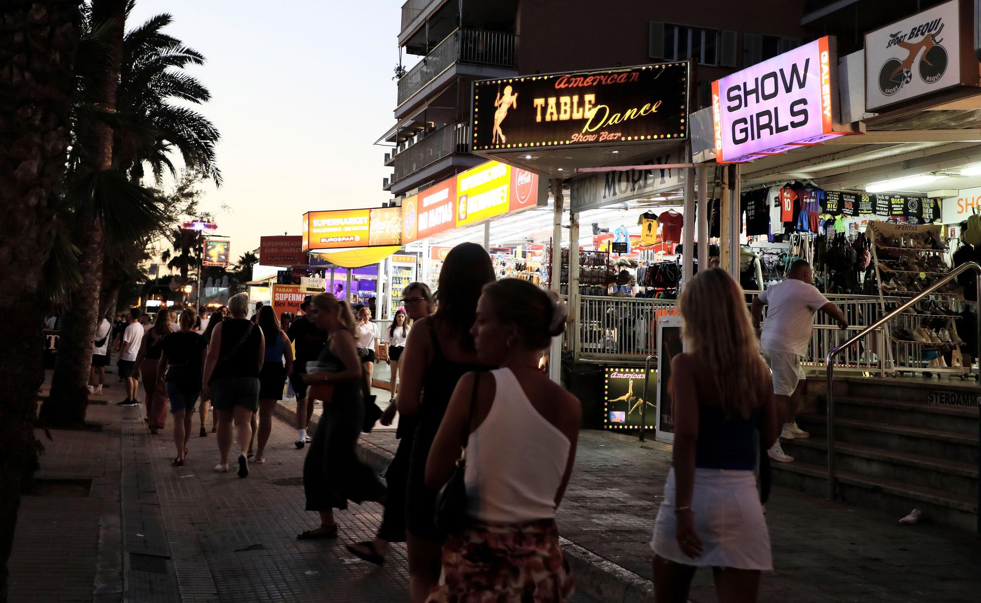 Menschen spazieren auf der Strandpromenade von El Arenal auf Mallorca an Show-Bars vorbei.