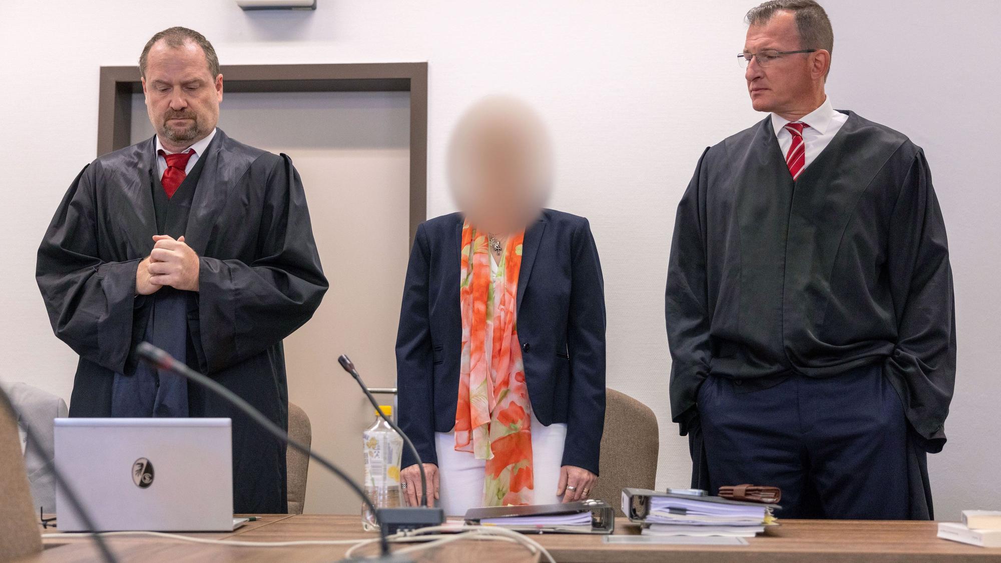 Eine Frau steht an der Anklagebank zwischen zwei Anwälten.