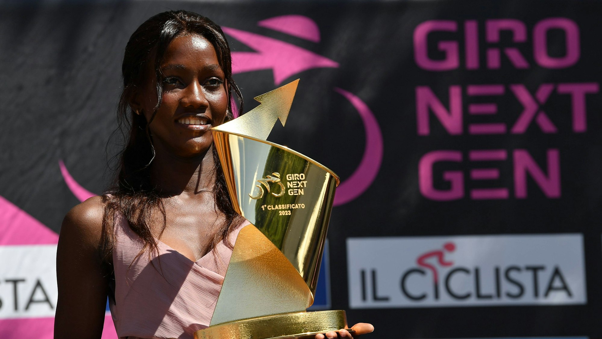 Eine Frau hält einen goldenen Pokal in den Händen. Im Hintergrund ist der Schriftzug Giro Next Gen zu sehen.