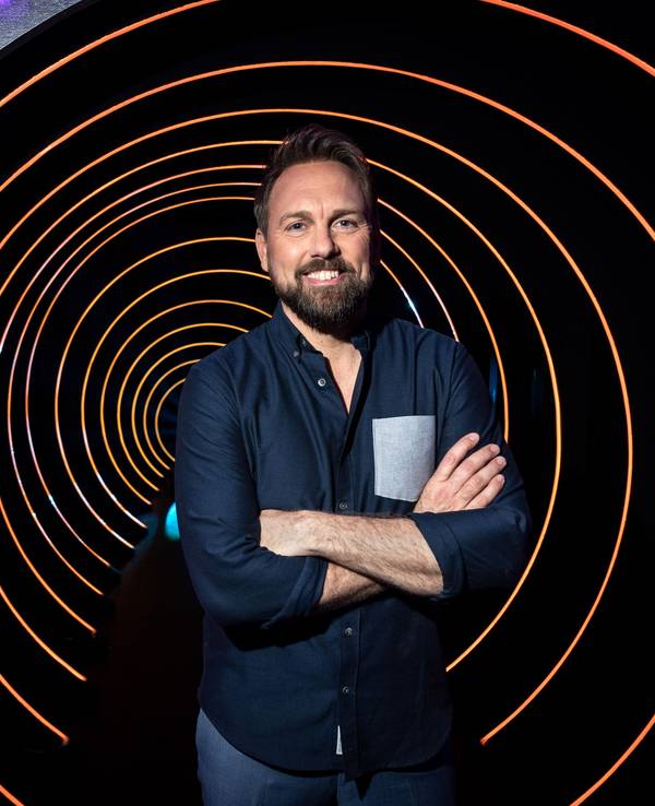 Moderator Steven Gätjen steht mit verschränkten Armen in der Kulisse der ProSieben-Sendung „Die Herzschlagshow“ und lächelt.