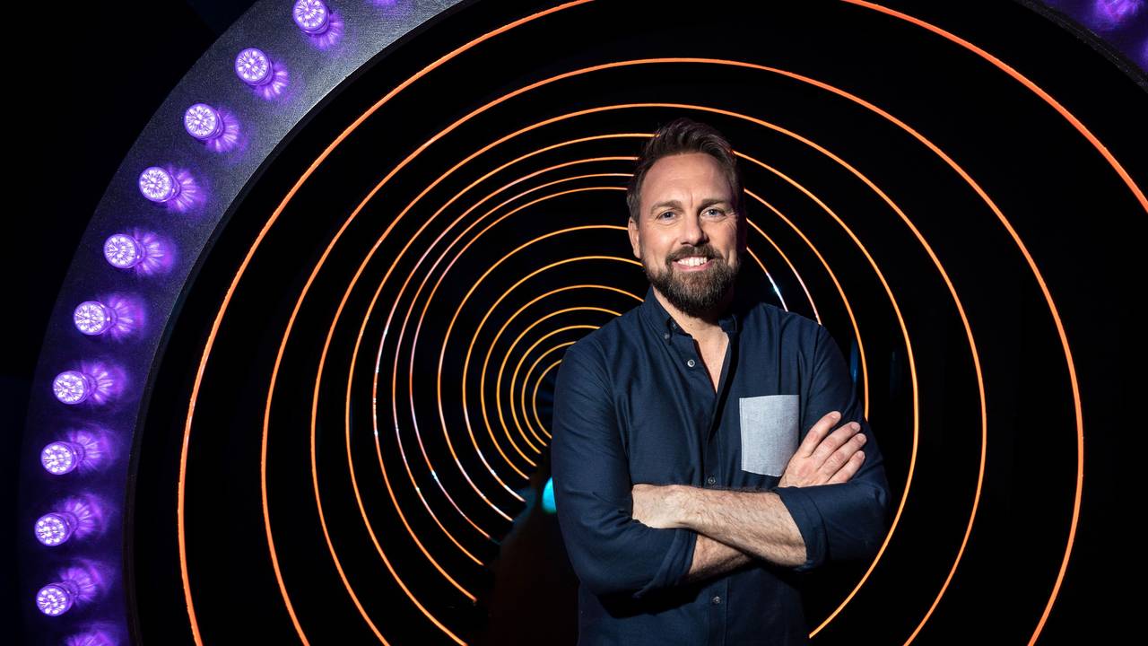 Moderator Steven Gätjen steht mit verschränkten Armen in der Kulisse der ProSieben-Sendung „Die Herzschlagshow“ und lächelt.