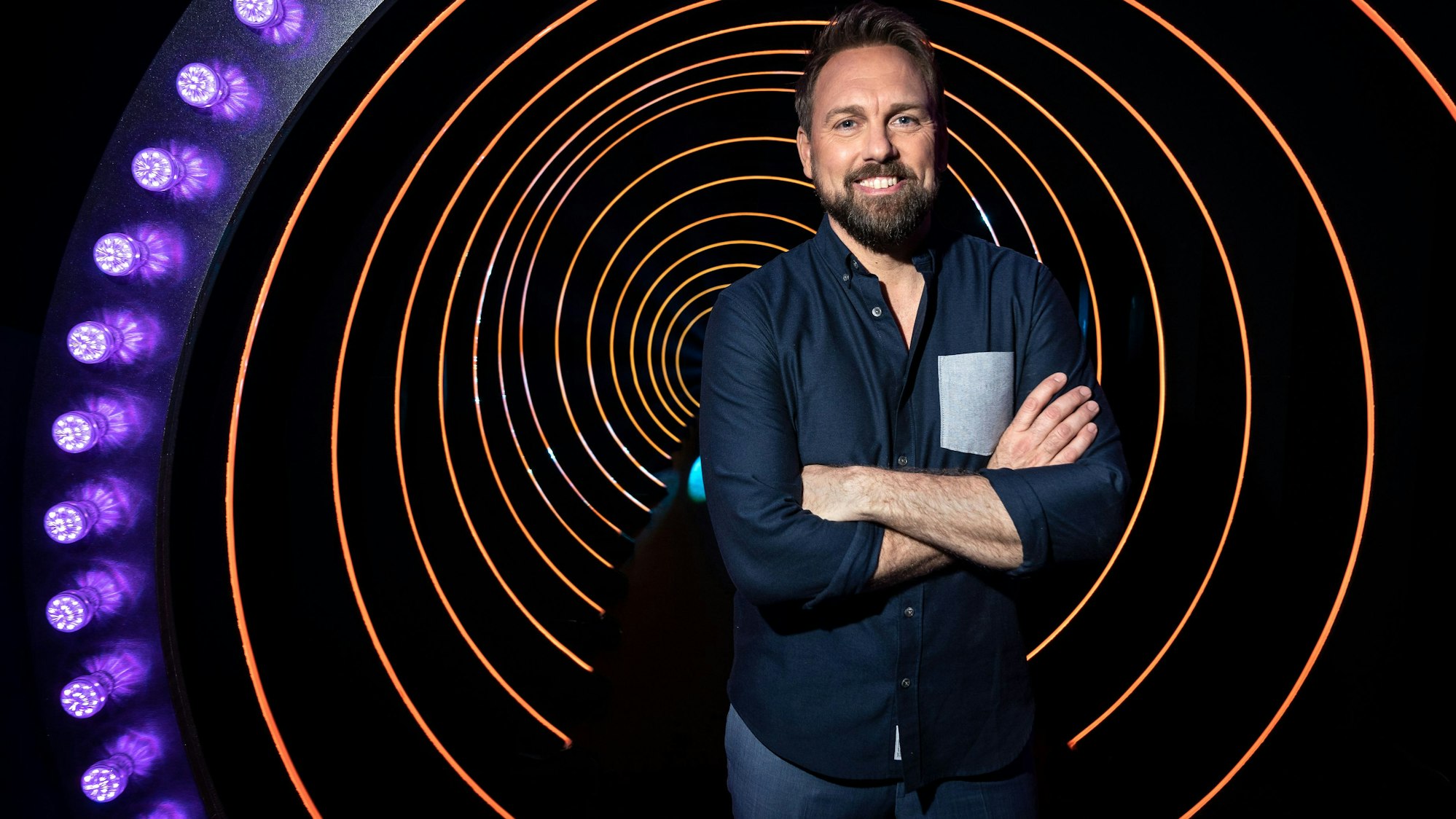 Moderator Steven Gätjen steht mit verschränkten Armen in der Kulisse der ProSieben-Sendung „Die Herzschlagshow“ und lächelt.