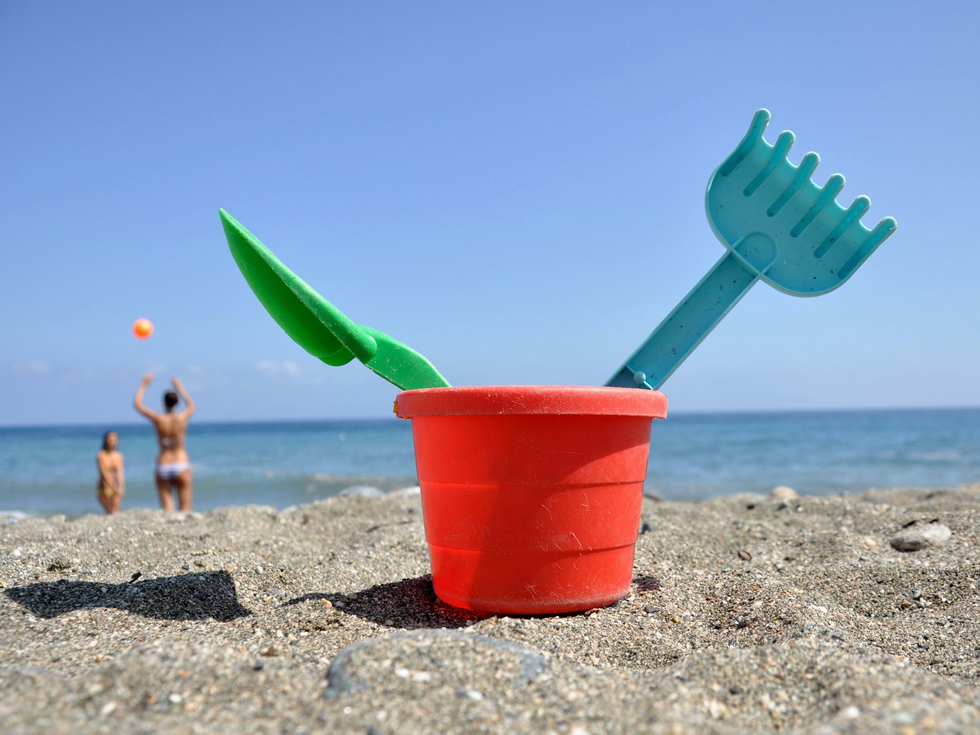 Strandspielzeug steht im Sand, im Hintergrund spielen zwei Frauen Volleyball.