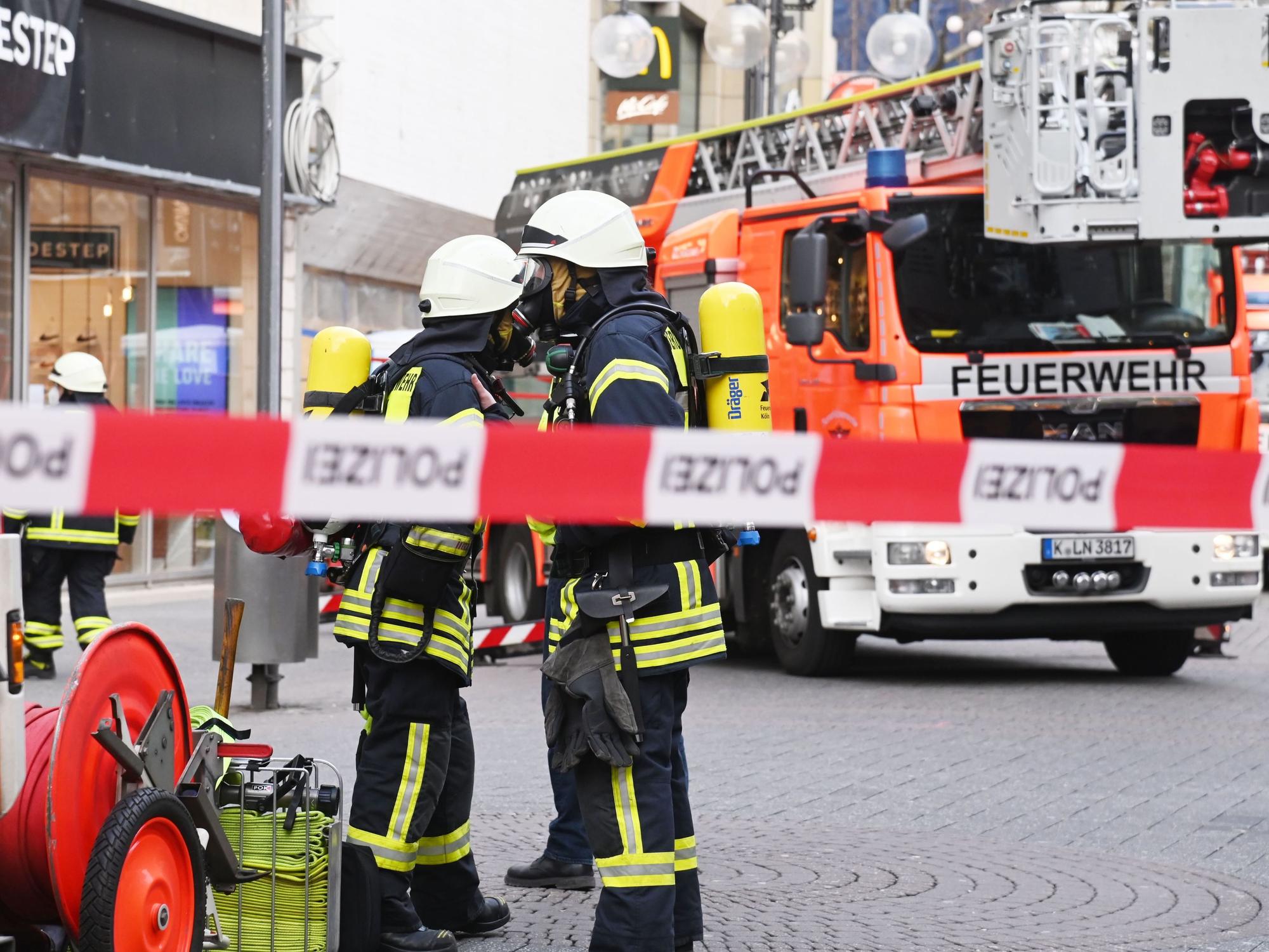 Feuerwehrleute und ein Feuerwehr-Auto stehen auf der Schildergasse in Köln. Im Vordergrund ist Absperrband zu sehen.