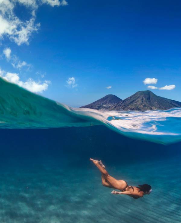 Eine Frau taucht im Meer, über ihr ist eine Insel mit zwei Vulkanen zu sehen.